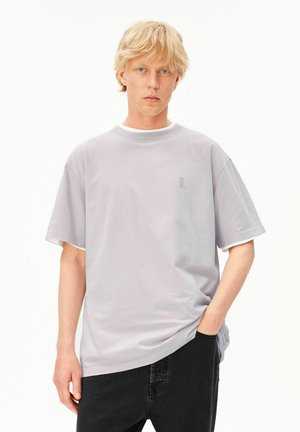 Blonder Mann mit hellen Augen, der ein lockeres hellgraues T-Shirt über einem weißen Hemd und schwarzen Hosen trägt, stehend mit einer Hand in der Tasche.