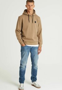 Felpa marrone con cappuccio e cordini, con un piccolo logo nero, abbinata a jeans azzurri e sneakers grigie. Tessuto morbido, vestibilità casual.