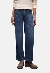 Vida jeans i mörk tvätt med vida ben, fyra fickor, toppsöm och avsmalnande fåll, i kombination med beige ankellånga stövlar.