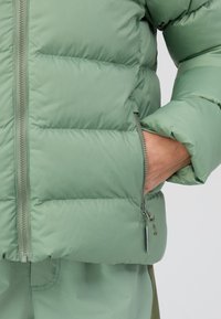 Gevoerde groene jas met ritsclosure, voorzien van een zijzak en zachte textuur. Inclusief zichtbare stiksels en een gladde stofafwerking.