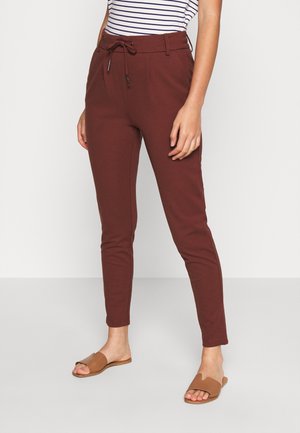 Vrouw draagt strakke bordeauxrode broek met een trekkoord in de taille, gecombineerd met bruine platte slippers en een gestreept topje.