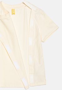 Kremowa koszula z krótkim rękawem, z przodu zapinana na szerokie białe rzepy dla łatwego ubierania.