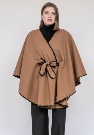 Femme portant une cape beige à bordure noire, nouée à la taille par un nœud, sur un col roulé noir et un pantalon noirs, debout devant un fond uni.