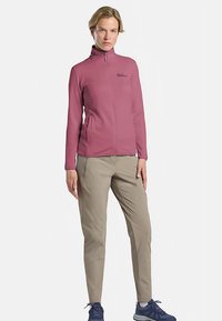 Veste zippée rose clair avec poches sur les côtés, associée à un pantalon beige ajusté et des chaussures de sport bleues, idéale pour les activités en plein air.