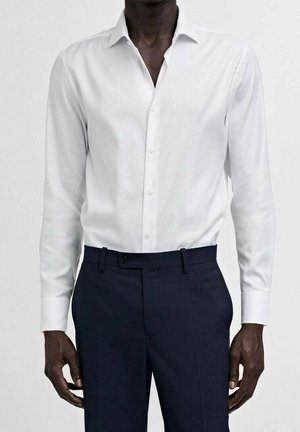 Homme portant une chemise blanche à boutons à manches longues et un pantalon ajusté bleu marine foncé, debout devant un fond clair.
