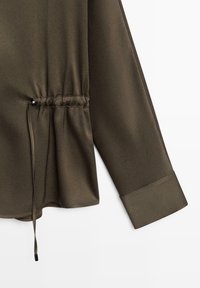 Blouse vert olive à manches longues, avec un détail de taille froncée et des manches à poignets. Tissu lisse et léger avec un éclat subtil.