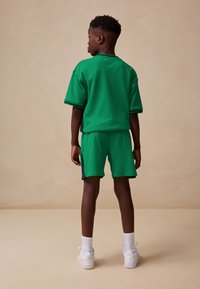 Green cotton blend athletic outfit featuring short sleeves, matching shorts, and accents in black. Worn with white sneakers and socks.