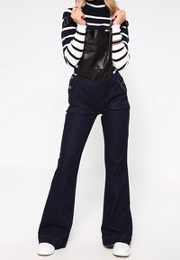 Zwarte leren overall met een brede tailleband en uitlopende donkerdenim benen, met een gestreepte lange mouwtop en witte sneakers.