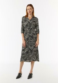 Czarna i biała sukienka w falistym wzorze z paskiem w talii, rękawami do trzech kwart, o zwiewnym kroju, zestawiona z czarnymi klapkami na obcasie.