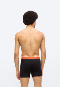 Μαύρα ανδρικά boxer briefs με κόκκινη ζώνη μέσης που φέρει το κείμενο "TOMMY HILFIGER". Λείο ύφασμα και εφαρμοστή σχεδίαση, εμφανιζόμενα από πίσω.