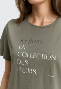 Olivgrünes Baumwoll-T-Shirt mit kurzen Ärmeln und weißem Text: „les fleurs LA COLLECTION DES FLEURS.“ Markenlogo „TOM TAILOR“ am Saum.