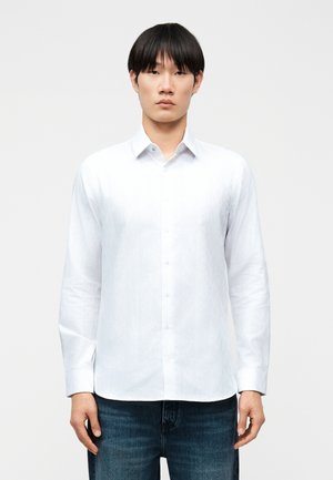 Mladý muž s rovnými čiernymi vlasmi, oblečený v jednoduchú bielu košeľu na gombíky a tmavomodré džínsy, stojaci pred bielym pozadím.