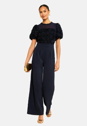 Femeie în pantaloni largi bleumarin și top texturat cu mâneci bufante, ținând o plic auriu, purtând pantofi aurii cu vârful deschis și cercei evidențiați.