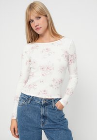 Weißes Langarmoberteil mit gerippter Textur und pastellfarbigem Blumenmuster. Kombiniert mit hochgeschnittenen Jeans, die über Taschen verfügen und eine lockere Passform bieten.