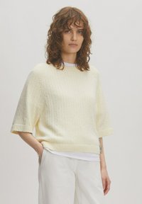 Cremfarbene, gestrickte Bluse mit kurzen Ärmeln, strukturierter Oberfläche und rundem Ausschnitt, kombiniert mit weißen, weit geschnittenen Hosen.