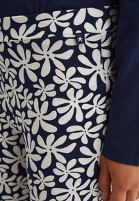 Sötétkék anyag fehér virágmintával; kis csillagcímke található rajta; sima tapintású; közeli felvétel az anyagról és mellette egy kéz.