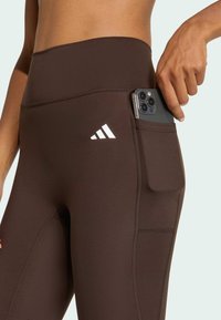 Leggings de sport marron avec une poche latérale contenant un smartphone. Présente un logo blanc à trois bandes sur la cuisse gauche. Texture lisse, design ajusté.