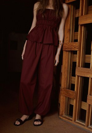 Donna con top smanicato rosso scuro a peplo e pantaloni larghi abbinati, in piedi vicino a una porta di legno all'interno.