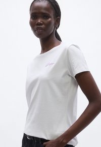 Fehér pamut póló kerek nyakkal és rövid ujjú. Elöl apró, világoslila hímzett logó díszíti. Sima tapintású anyag.