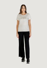 Giovane donna che indossa una t-shirt bianca a maniche corte con logo, pantaloni larghi neri e scarpe stringate nere, in piedi contro uno sfondo chiaro.