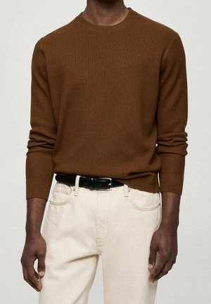 Homme portant un pull à manches longues brun côtelé rentré dans un pantalon écru avec une ceinture noire, debout les bras détendus le long du corps.