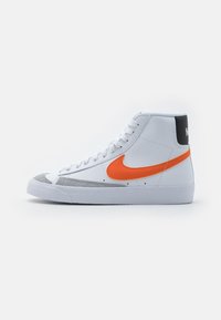 Vit högsko med grå tåhuvud, orange Nike-logga, vita snören och svart bakparti på en enkel bakgrund.