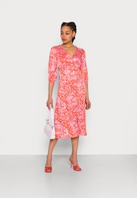 Vestido com padrão em vermelho e rosa, com decote em V, mangas bufantes até ao cotovelo e detalhes em botão. Combinado com uma mala rosa clara e saltos vermelhos.