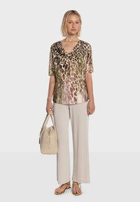 Haut à manches courtes et col bénitier avec un imprimé léopard aux tons verts et roses, associé à un pantalon beige à jambes larges et des tongs dorées, tenant un sac à main beige.