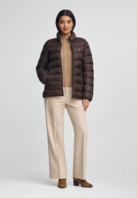 Brun vadderad jacka med hög krage, bärs över en beige kabelstickad tröja, tillsammans med beigefärgade byxor och bruna klackade stövlar vid ankeln.