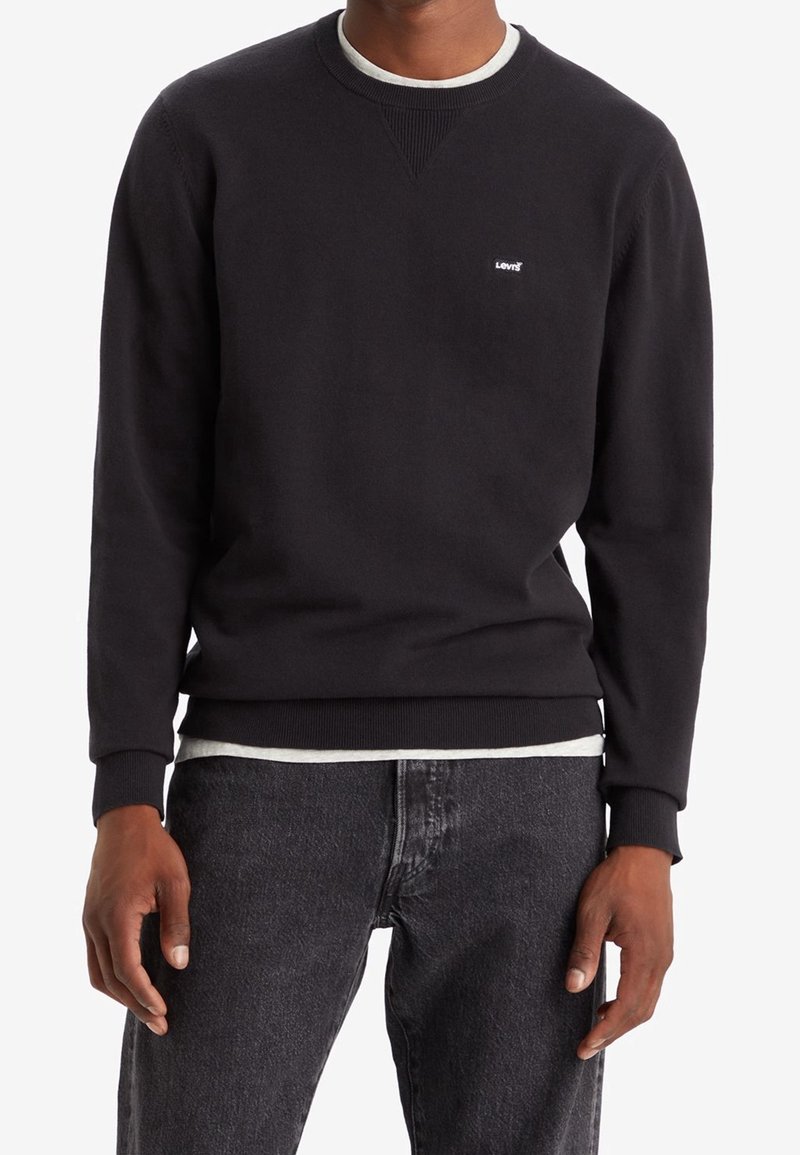 Homme portant un sweat-shirt col rond noir avec un petit logo blanc sur la poitrine, superposé à un T-shirt blanc et associé à un jean gris foncé.