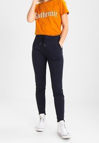 Camiseta de algodón naranja con el texto "Auténtico", combinada con pantalones joggers azul marino con bolsillos laterales y puños elásticos, usados con zapatillas blancas.