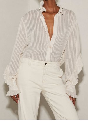 Femme portant un chemisier blanc transparent à manches longues avec des volants et des rayures verticales, rentré dans un pantalon blanc cassé taille haute.