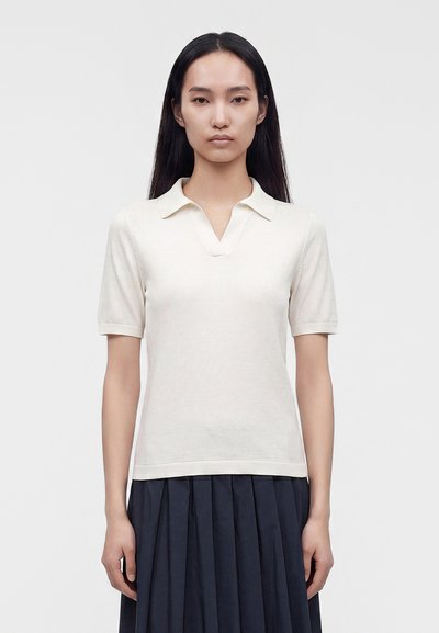 Femme aux longs cheveux noirs portant une chemise blanche à manches courtes avec col et une jupe plissée foncée, debout devant un fond clair uni.