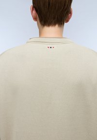 Persoon met kort bruin haar, gekleed in een lichtbeige trui, die de achterkant toont met een klein rood, wit en blauw geborduurd logo nabij de kraag.