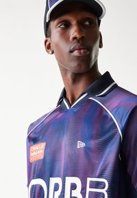 Πόλο πουκάμισο με ναυτικό μπλε και μοβ κυματιστό σχέδιο, λευκές λεπτομέρειες και λογότυπο. Το μοντέλο φοράει ένα καπέλο που ταιριάζει με δομημένο γείσο.