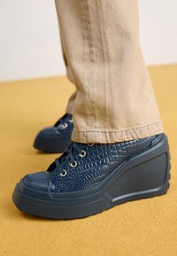 Námornické wedge tenisky s embosovaným textilním vzorem krokodýla, gumovým špičkovým krytem, zlatými očky a texturovanou podrážkou, spárované se světle béžovými kalhotami.