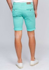 Shorts turquoise en tissu texturé, avec ourlets roulés, deux poches arrière avec fermetures à bouton, et une étiquette de marque en cuir marron au niveau de la taille.