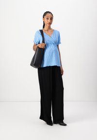 Hellbluse mit Lochmuster und kurzen Rüschensäumen, kombiniert mit lockeren schwarzen Hosen. Trägt eine schwarze Tote-Tasche.
