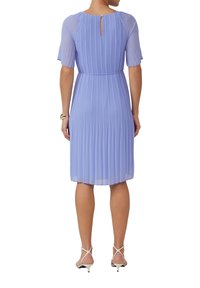 Plissiertes Kleid in Helllila mit kurzen Ärmeln und einem Schlüsselelement im Rücken. Getragen mit weißen Riemchenschuhen. Geschmeidiger, fließender Stoff.