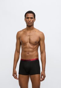 Boxer noir avec une ceinture élastique rouge arborant le logo "HUGO". Tissu lisse, coupe ajustée et longueur de jambe standard.