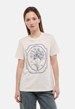 Femme portant un T-shirt crème à manches courtes avec une illustration florale encadrée bleu marine intitulée « Agapanthe » et un jean bleu.