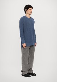 Pullover blu a coste con scollatura ampia e maniche lunghe, abbinato a pantaloni grigi a gamba larga e scarpe nere. Nessun motivo visibile.