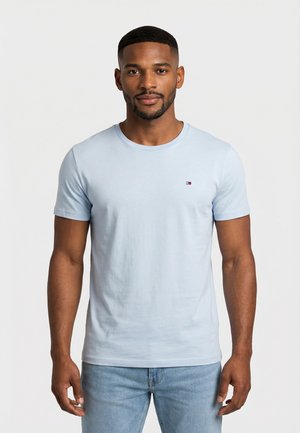 Man wearing light blue short-sleeve T-shirt with small logo on chest and light blue jeans, standing against a plain white background.