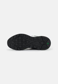 Suela de zapatilla de goma negra con una superficie texturizada y patrones de relieve multifuncionales, logo identificativo y detalle en color verde.