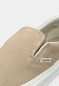 Chaussure beige à enfiler avec dessus en daim, semelle blanche, panneaux latéraux élastiques et petite étiquette en tissu portant la marque Hilfiger près du col.