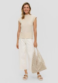 Beige textuur bovenstuk met een gegolfde halslijn, gecombineerd met brede crèmekleurige broek en een geweven schoudertas. Model draagt lichte sandalen.