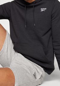 Sweat à capuche noir avec cordon de serrage réglable et poche avant, arborant un petit logo Reebok blanc. Associé à un short gris.