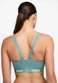 Mujer con cabello castaño en una coleta, lleva un sujetador deportivo Nike de color azul claro, vista desde atrás sobre un fondo blanco liso.