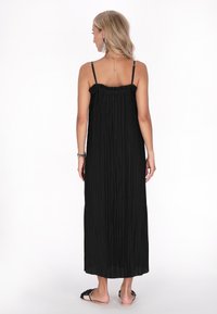 Schwarzes, plissiertes Maxikleid mit dünnen, verstellbaren Trägern und einem rüschierten Ausschnitt. Glatte Stoffdrapierung bis zur Knöchellänge, kombiniert mit Slipper-Sandalen.