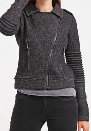 Vrouw draagt een gestructureerd donkergrijs jack met ritssluiting en geribbelde mouwen, gecombineerd met een grijze blouse en zwarte broek.